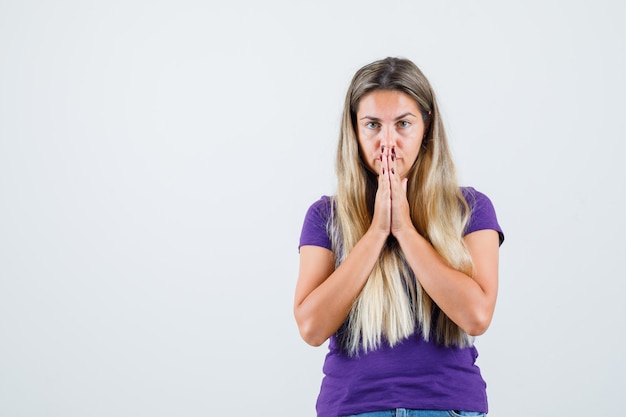 Blonde lady holding hands in praying gesture in violet t-shirt and looking calm , front view.