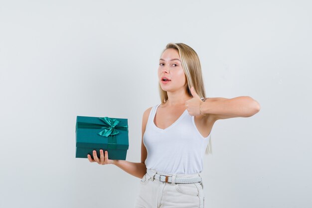 Blonde lady holding present box, showing thumb up in singlet, pants and looking confident. front view.