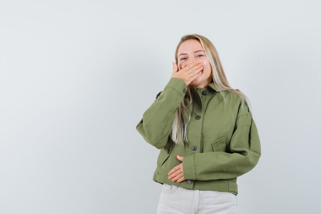 Blonde lady in jacket, pants holding hand on mouth and looking happy , front view.