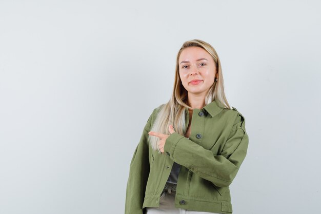 Blonde lady in jacket, pants pointing aside and looking confident , front view.