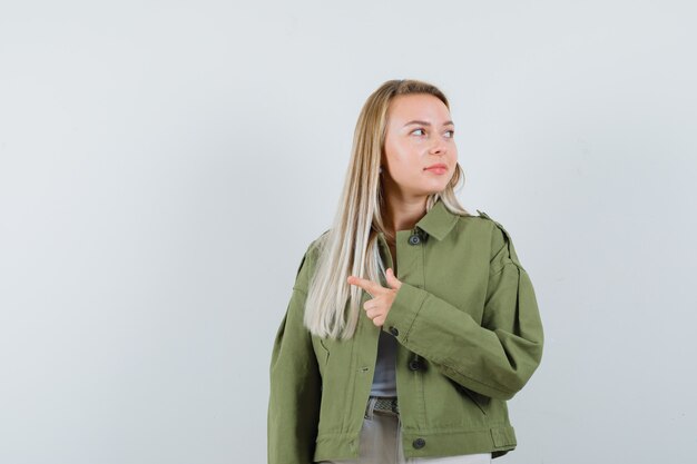 Blonde lady in jacket, pants pointing aside and looking focused , front view.