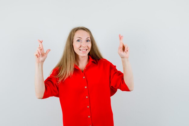 Blonde lady keeping fingers crossed in red shirt and looking cheerful.