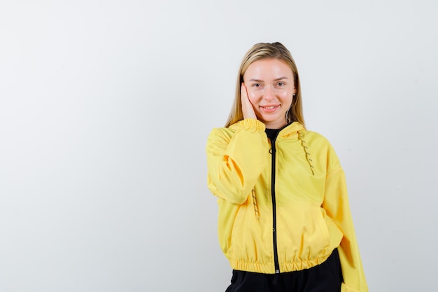 Blonde lady keeping hand on cheek in tracksuit and looking cheery. front view.