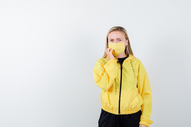 Blonde lady keeping hand on cheek in tracksuit, mask and looking anxious , front view.