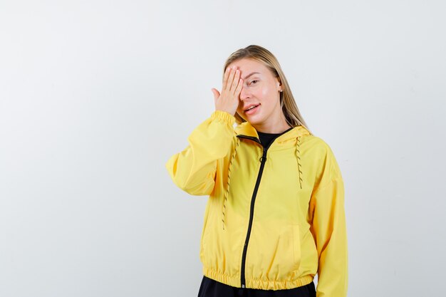 Blonde lady keeping hand on eye in tracksuit and looking cheerful. front view.