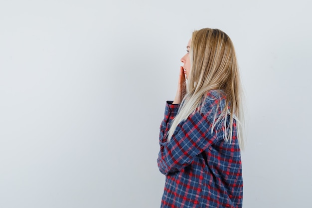 Blonde lady keeping hand on mouth in casual shirt and looking surprised .