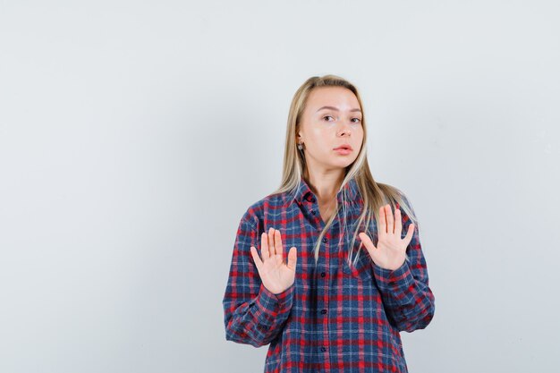 Blonde lady keeping hands to defend herself in casual shirt and looking careful. front view.