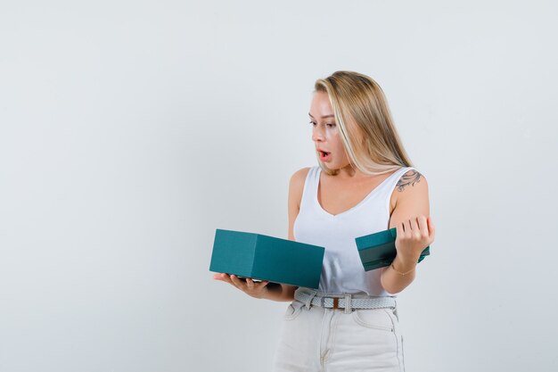 Blonde lady looking into present box in singlet, pants and looking shocked , front view.