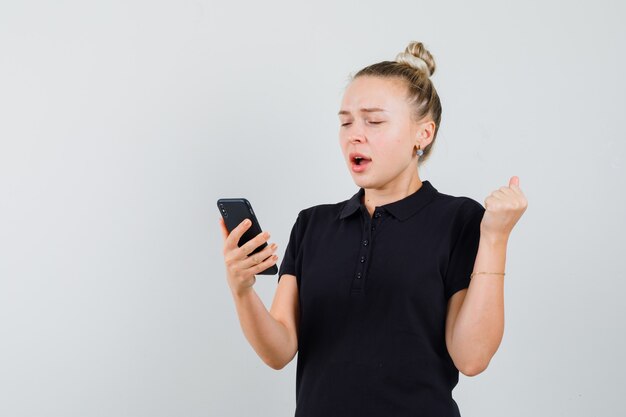 携帯電話を見て、黒のTシャツ、正面図で勝者のジェスチャーを示す金髪の女性。