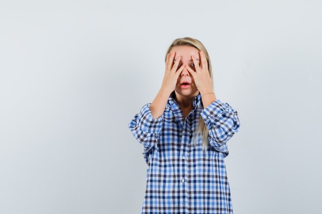 Blonde lady looking through fingers in checked shirt and looking scared