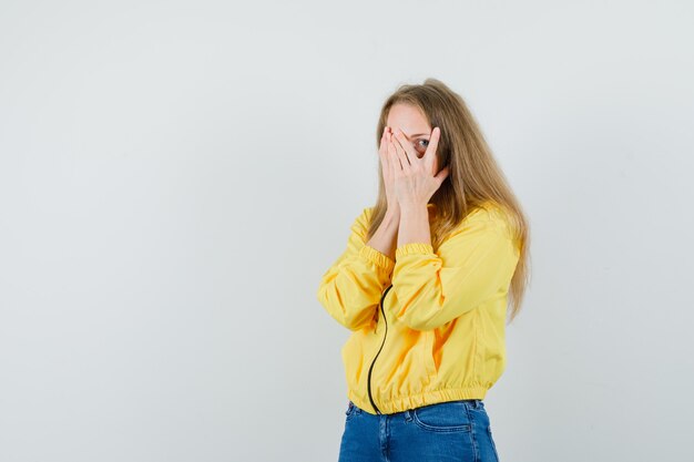 Blonde lady looking through fingers in jacket, jeans and looking scared.