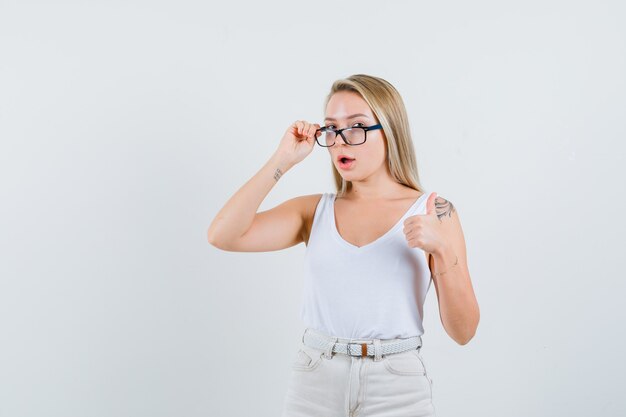 Blonde lady looking through glasses, showing thumb up in singlet, pants , front view.