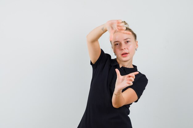 Blonde lady making frame gesture in black t-shirt and looking confident. front view.