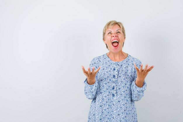 Blonde lady in patterned blouse raising hands with open palm while screaming and looking energetic , front view. free space for your text