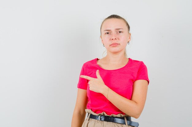 Blonde lady in pink t-shirt, pants pointing to the side and looking discontent , front view.