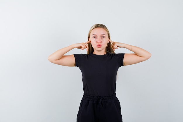 Blonde lady plugging ears with fingers, pouting lips in black dress and looking careful. front view.