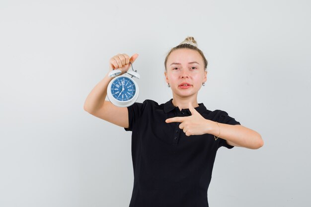 Blonde lady pointing at alarm clock in black t-shirt and looking confident. front view.