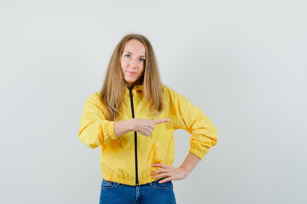 Blonde lady pointing aside in jacket, jeans and looking cheery.