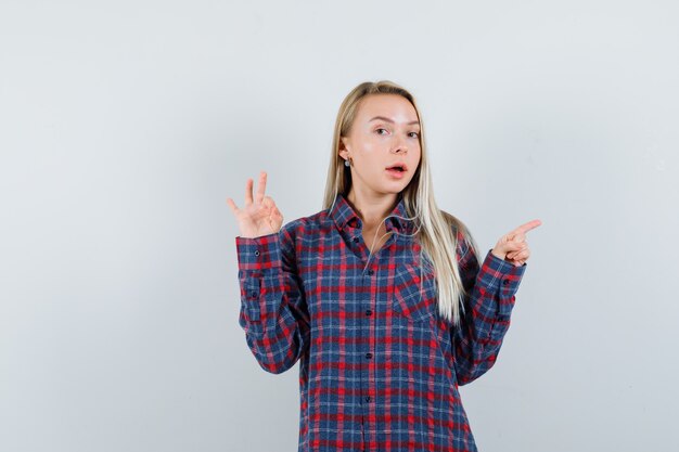 Blonde lady pointing aside, showing ok gesture in casual shirt front view.