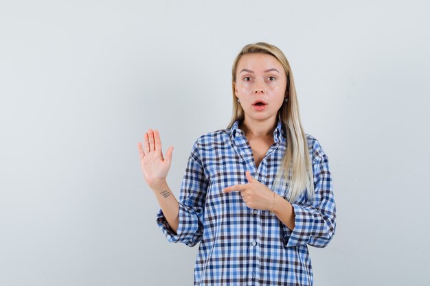 Blonde lady pointing aside, showing palm in checked shirt and looking wondered