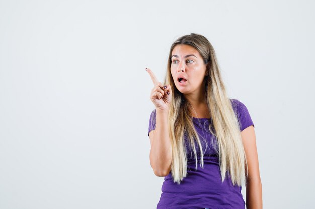 Blonde lady pointing away in violet t-shirt and looking surprised. front view.