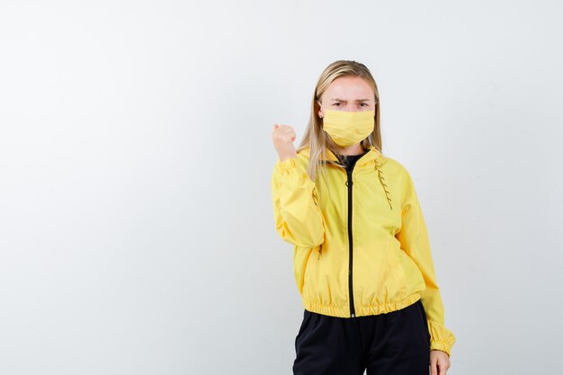 Blonde lady pointing back with thumb in tracksuit, mask and looking offended , front view.