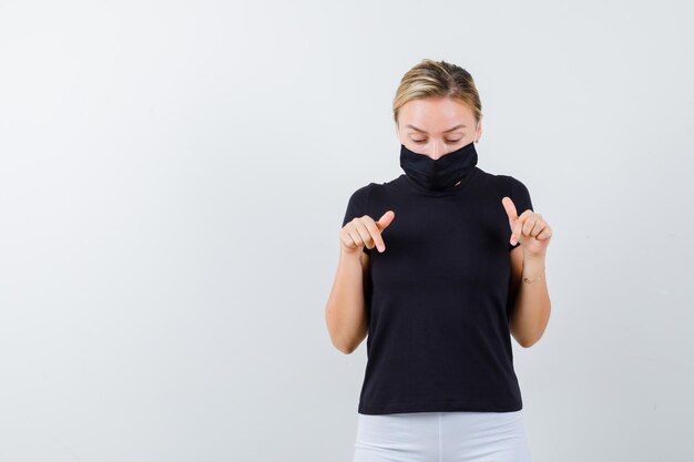 Blonde lady pointing down in black t-shirt, black mask and looking careful isolated