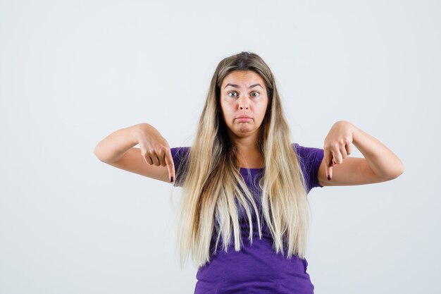 Blonde lady pointing down in violet t-shirt and looking puzzled , front view.