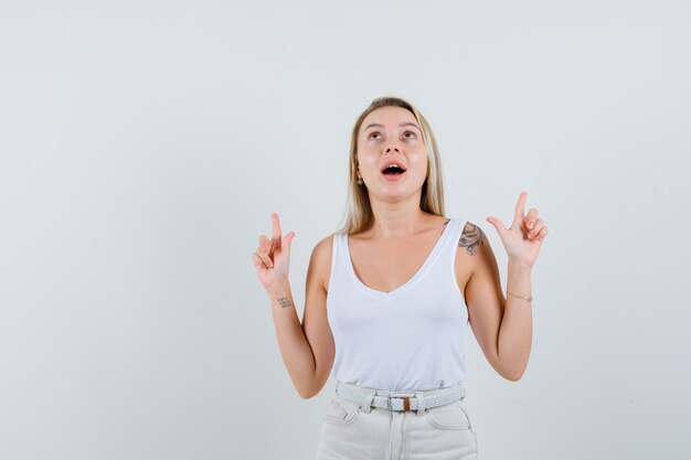 Blonde lady pointing fingers up in singlet, pants and looking grateful , front view.