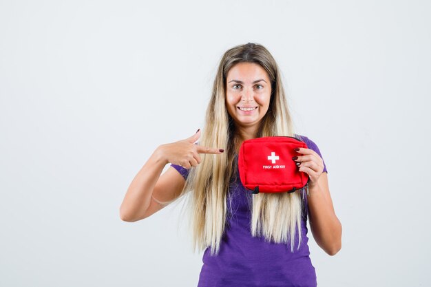 Blonde lady pointing at first aid kit in violet t-shirt and looking cheery , front view.