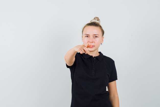 Blonde lady pointing at front in black t-shirt and looking confident , front view.