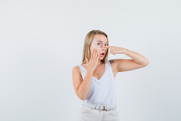 Blonde lady pointing at her eye in singlet, pants and looking cute