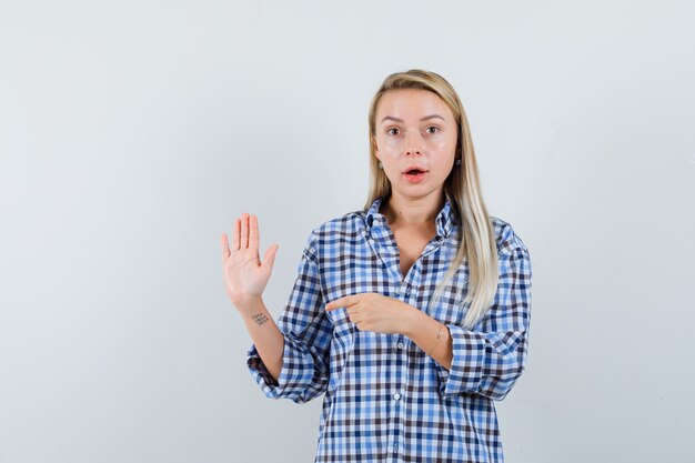 Blonde lady pointing at her raised palm in checked shirt and looking amazed