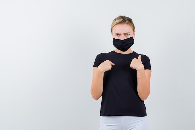 Blonde lady pointing at herself in black t-shirt, black mask and looking gloomy isolated