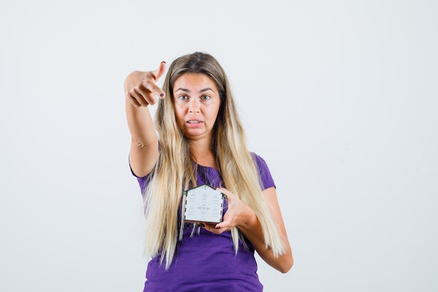 Blonde lady pointing at house model in violet t-shirt , front view.