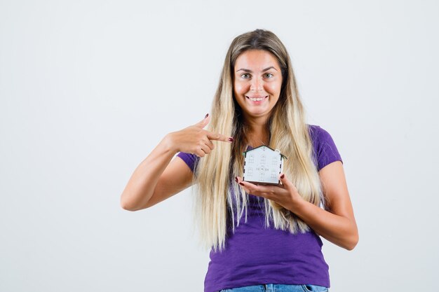 Blonde lady pointing at house model in violet t-shirt, jeans and looking cheerful , front view.