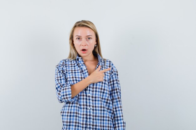 Blonde lady pointing to the right side in checked shirt and looking puzzled