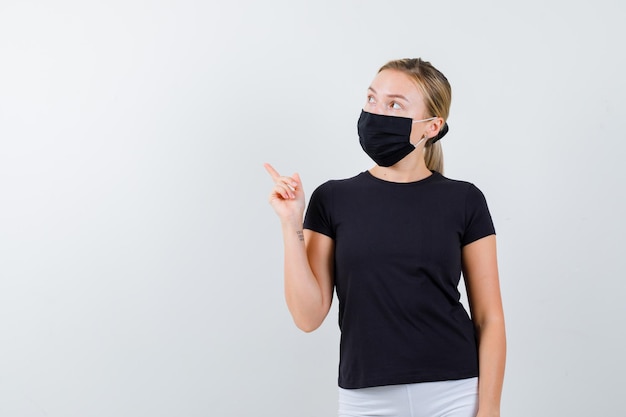 Blonde lady pointing up in black t-shirt, black mask and looking attentive isolated