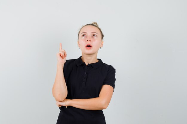 Blonde lady pointing up in black t-shirt and looking surprised. front view.