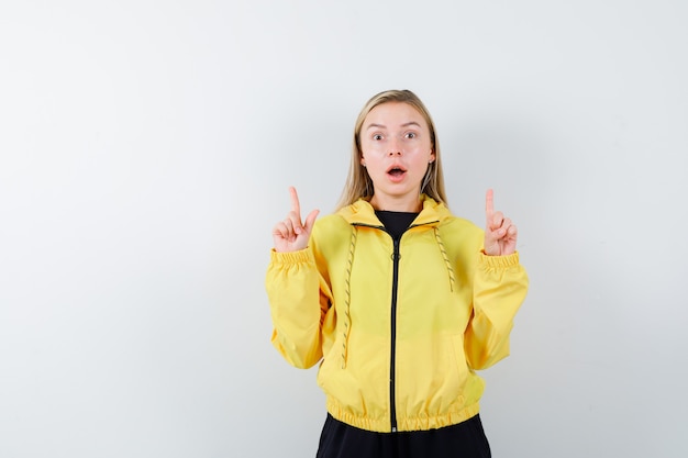 Blonde lady pointing up in tracksuit and looking shocked , front view.