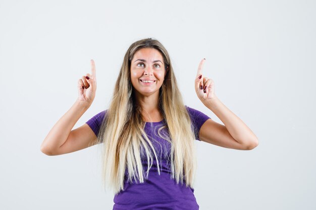 Blonde lady pointing up in violet t-shirt and looking joyful , front view.