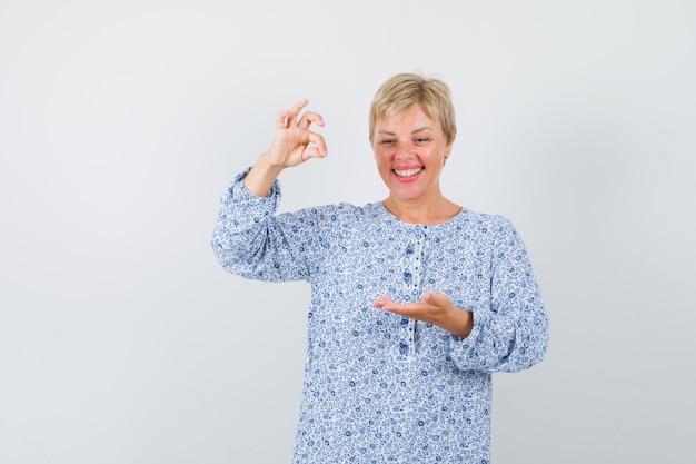 Blonde lady posing like pinching something in patterned blouse and looking joyous , front view.