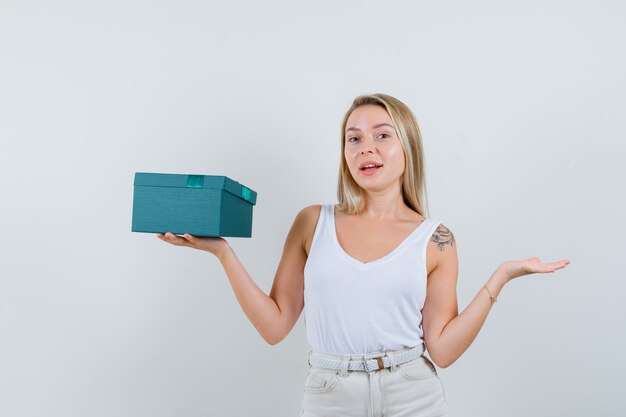 Blonde lady presenting present box in singlet, pants and looking jolly , front view.