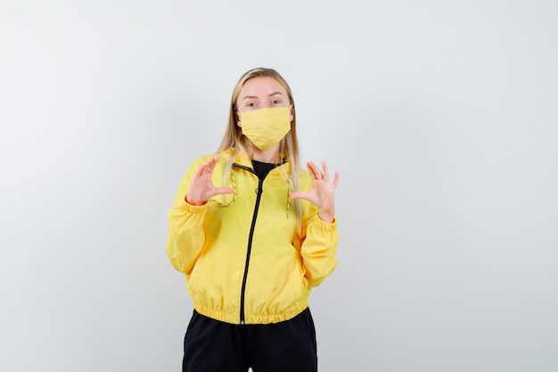 Free photo blonde lady pretending to hold or show something in tracksuit, mask and looking confident , front view.