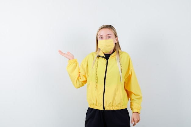 Blonde lady pretending to show something in tracksuit, mask and looking bewildered , front view.