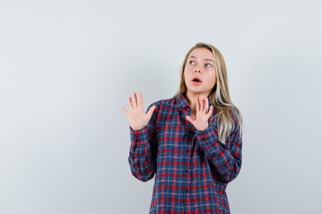 Blonde lady raising hands to defend herself in casual shirt and looking scared , front view.