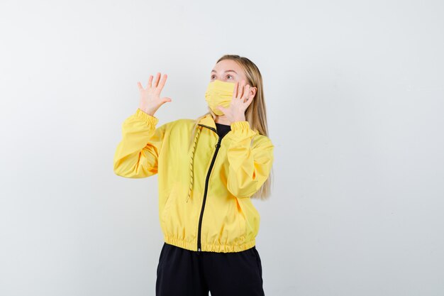 Blonde lady raising hands in surrender gesture in tracksuit, mask and looking scared. front view.