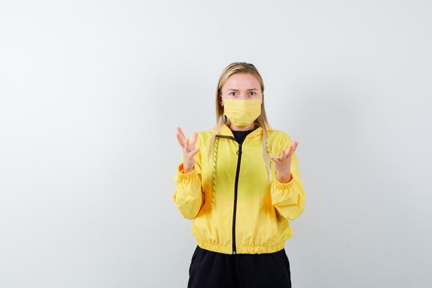 Blonde lady raising hands in tracksuit, mask and looking bewildered. front view.