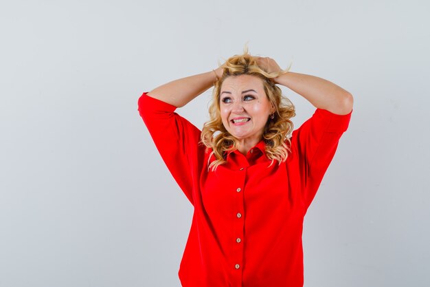 Blonde lady in red shirt holding hands on hair and looking amused
