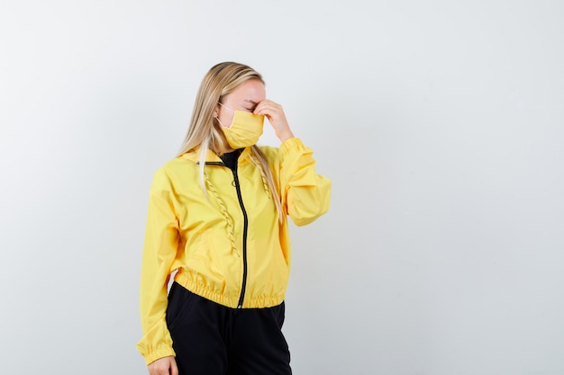 Free photo blonde lady rubbing eyes and nose in tracksuit, mask and looking tired , front view.